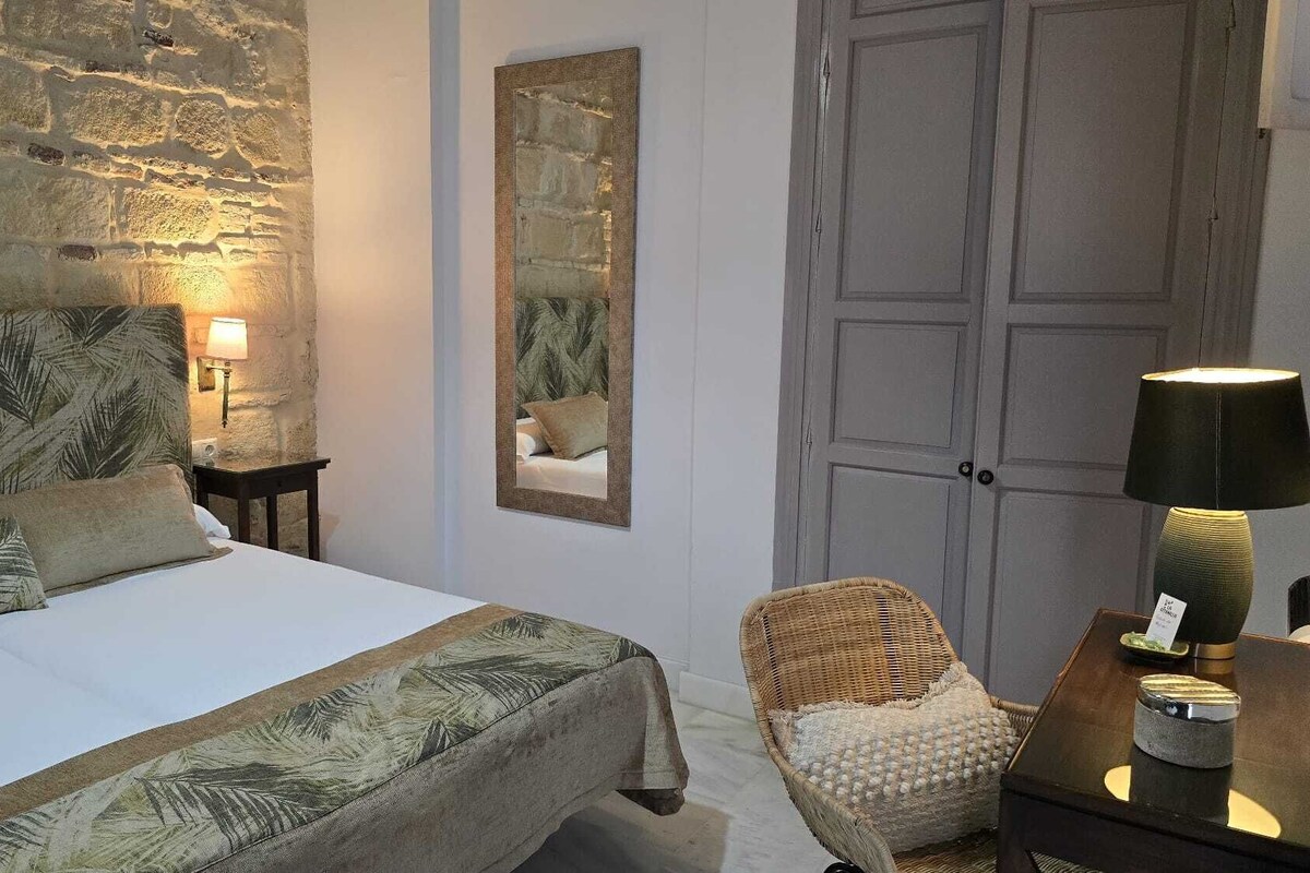 A bedroom with a stone wall, a bed with a leaf-patterned bedspread, a nightstand with a lamp, a wicker chair, and a door.