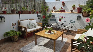 A cozy outdoor patio with a wooden table, a sofa, and two chairs. The area is adorned with various potted plants and flowers.