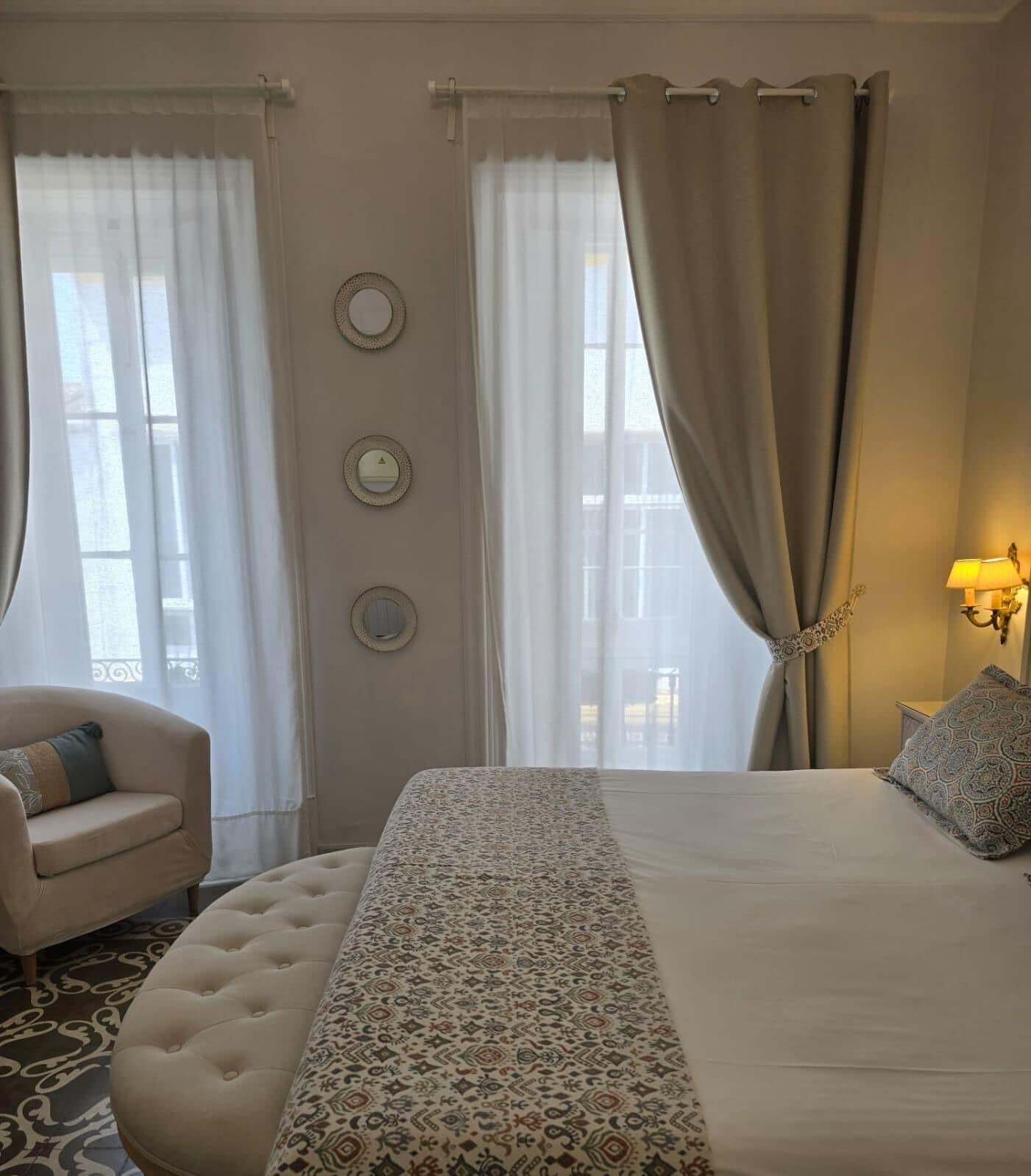 A hotel room with a bed, a chair, a lamp, and three wall-mounted mirrors.