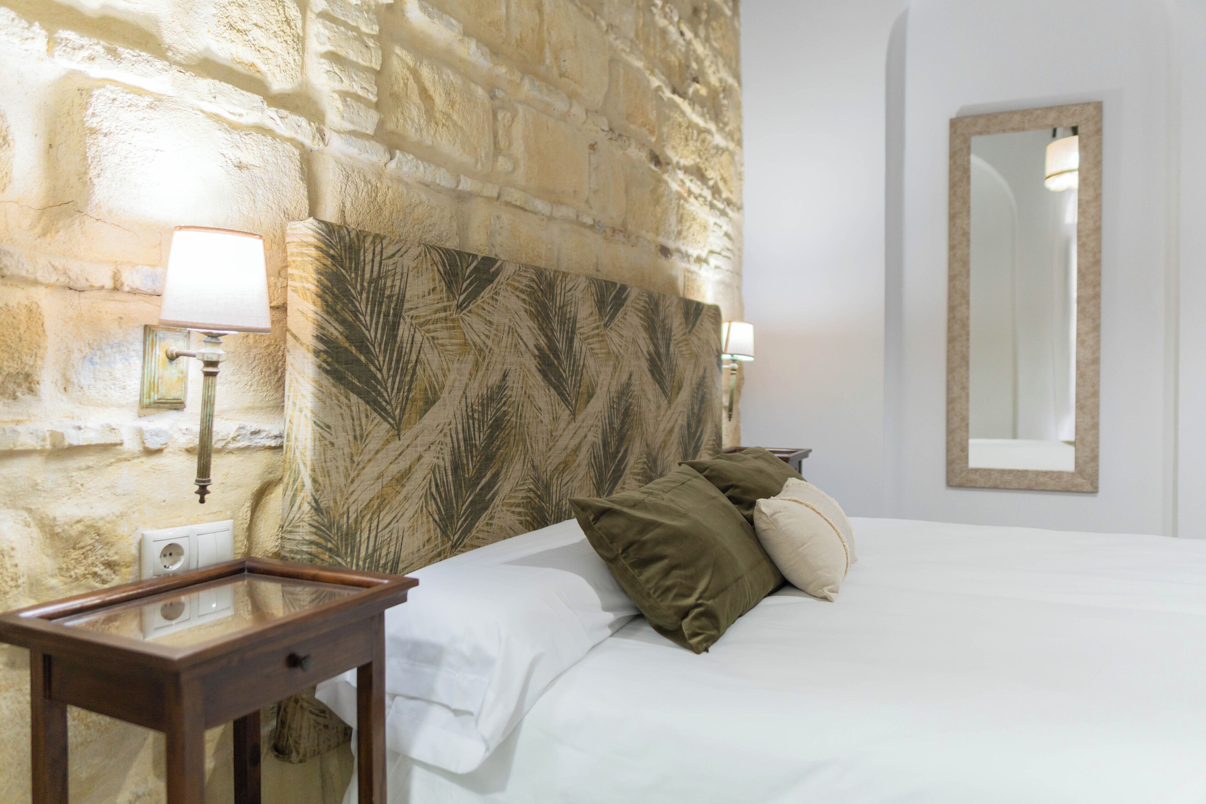 Habitación de hotel con pared de piedra, cama con ropa blanca, mesita de noche de madera, lámpara y espejo.