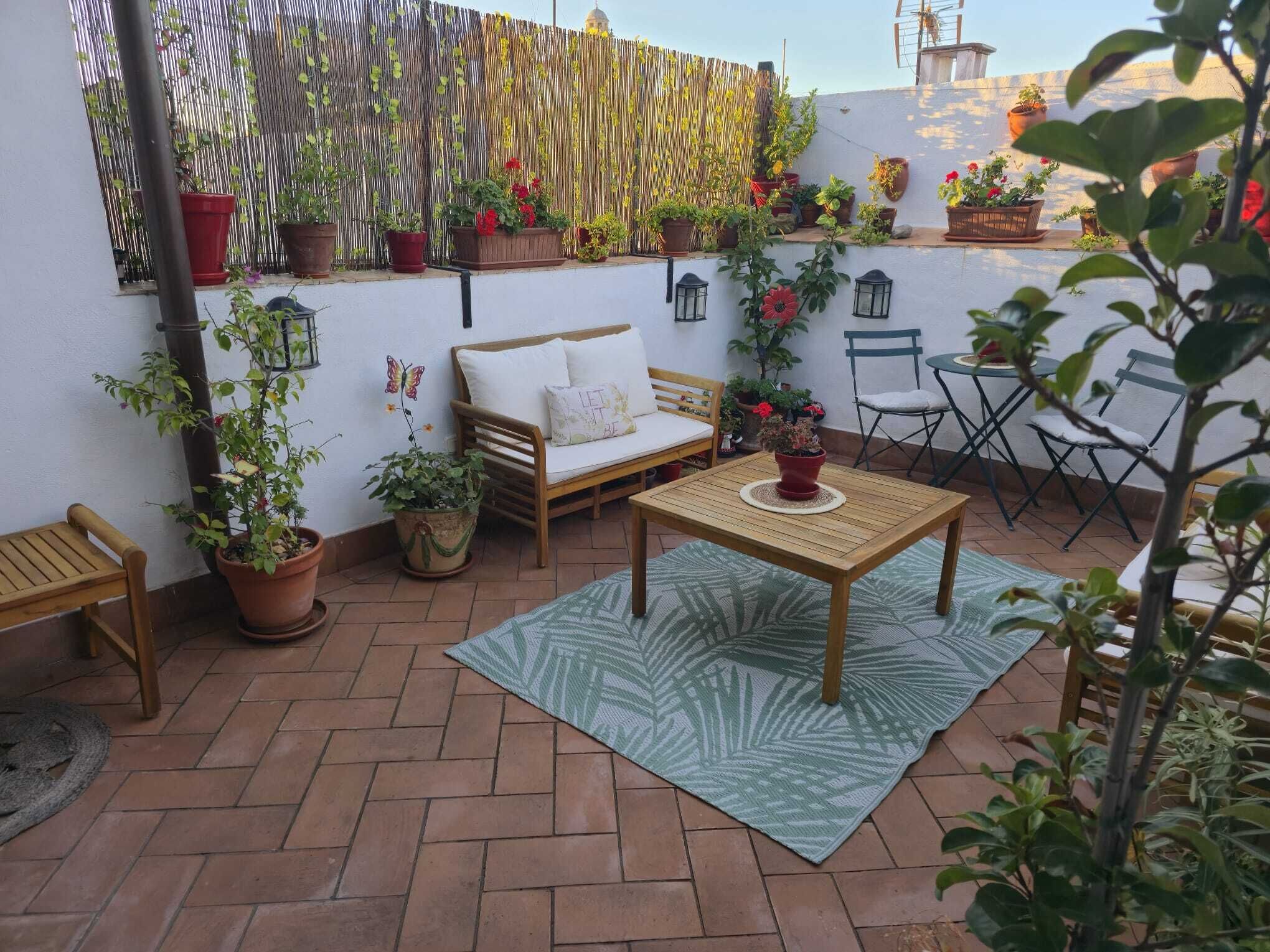 Une terrasse avec une table en bois, un canapé et des chaises, entourée de plantes en pot et d’une clôture en bambou.
