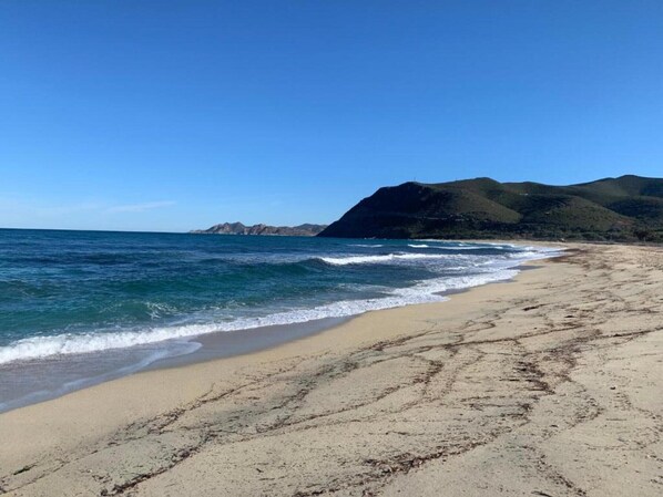 A beach with clear blue water, sandy shore, and green hills in the background. - Holiday Park in Corsica with Terrace Homes (Belgodere)