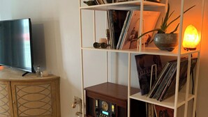 A living room with a white shelving unit filled with books, decorative items, and a small radio. A television is on a wooden cabinet to the left, and a guitar is on the floor.
