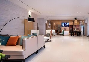 A modern hotel lobby with a seating area, a dining table, and a television screen.