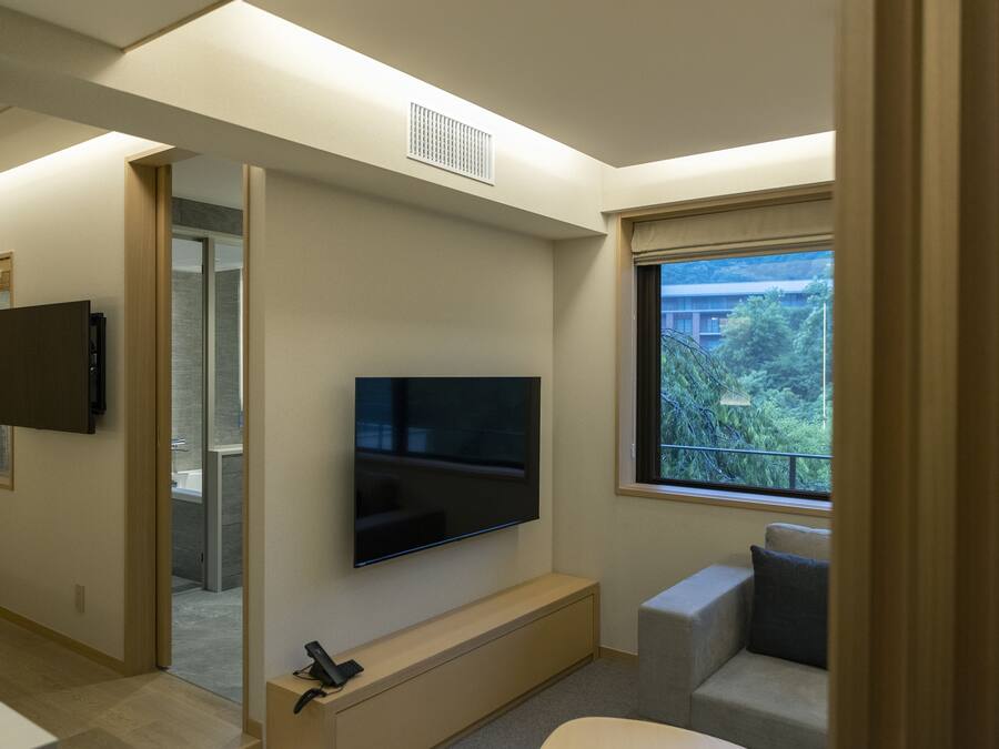 Ein modernes Wohnzimmer mit Flachbildfernseher an der Wand, einer Couch und einem Fenster mit Blick ins Grüne.