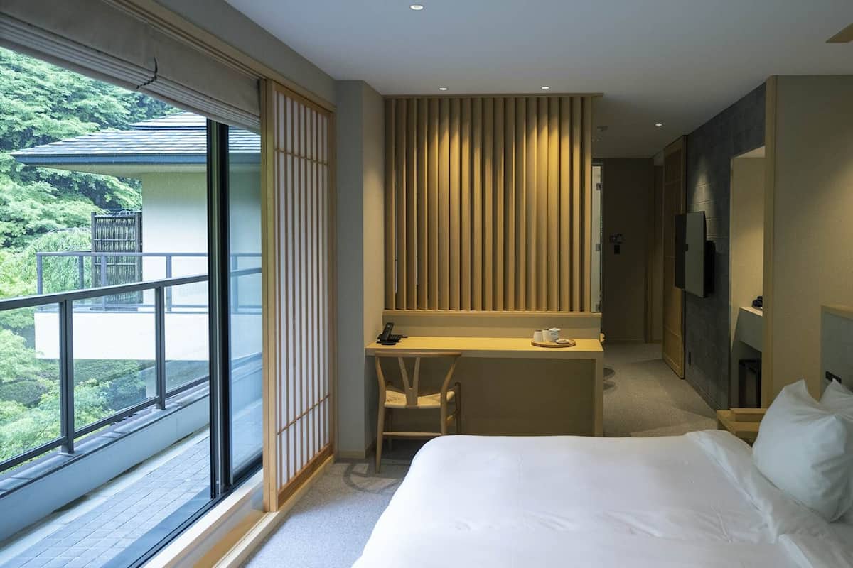 A hotel room with a bed, a desk, a TV, and a view of greenery outside.