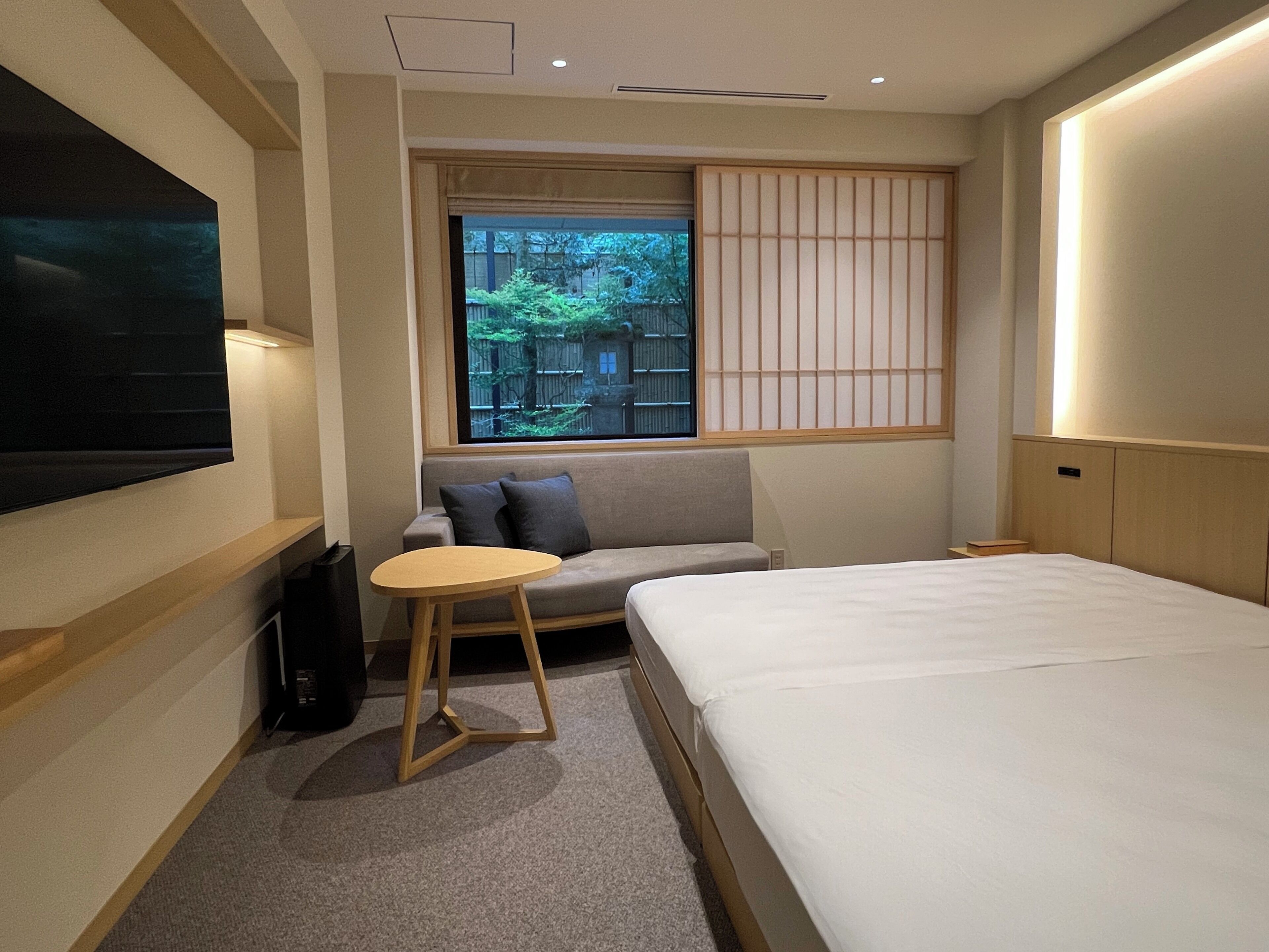 A hotel room with a bed, a sofa, a small round table, a TV mounted on the wall, and a window with a view of greenery.