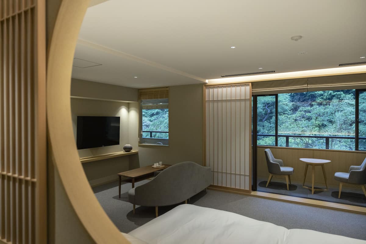 Ett modernt hotellrum med en säng, en TV, ett litet bord och två stolar nära ett fönster med utsikt över träd.