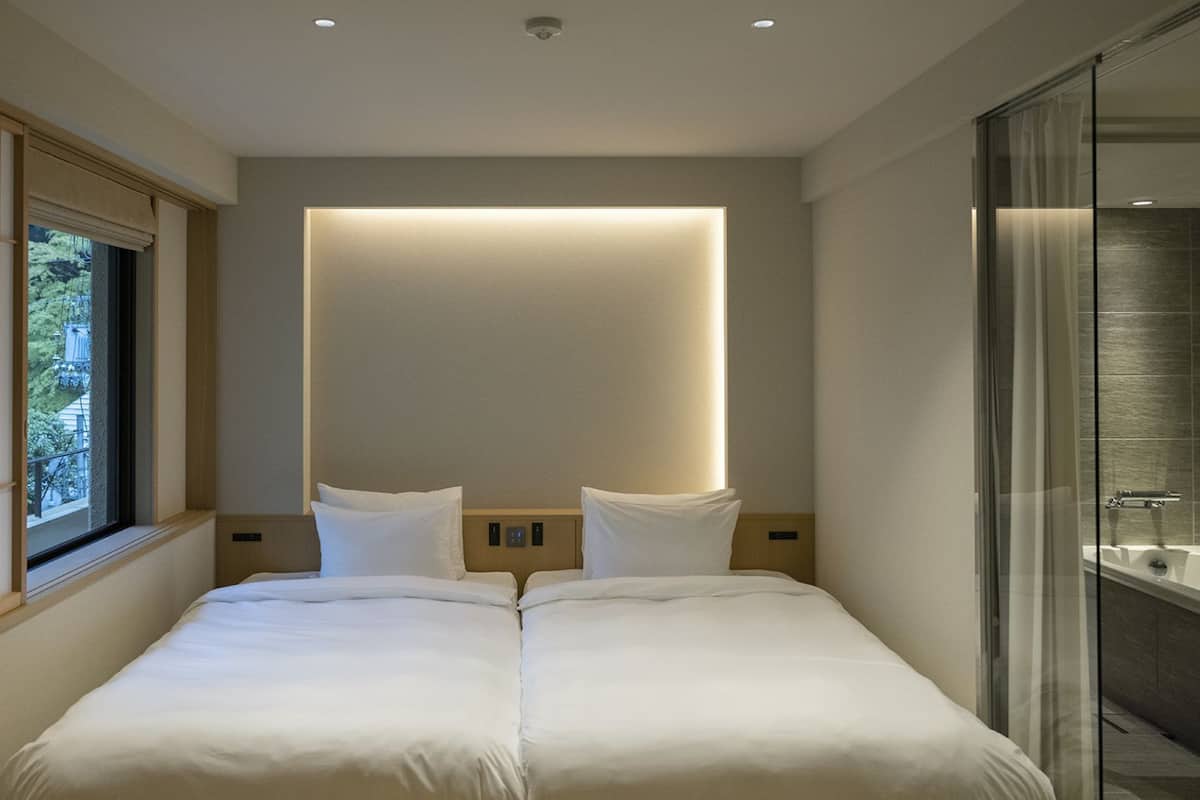 A modern hotel room with a large bed, a window with blinds, a bedside table, and a glass shower area.