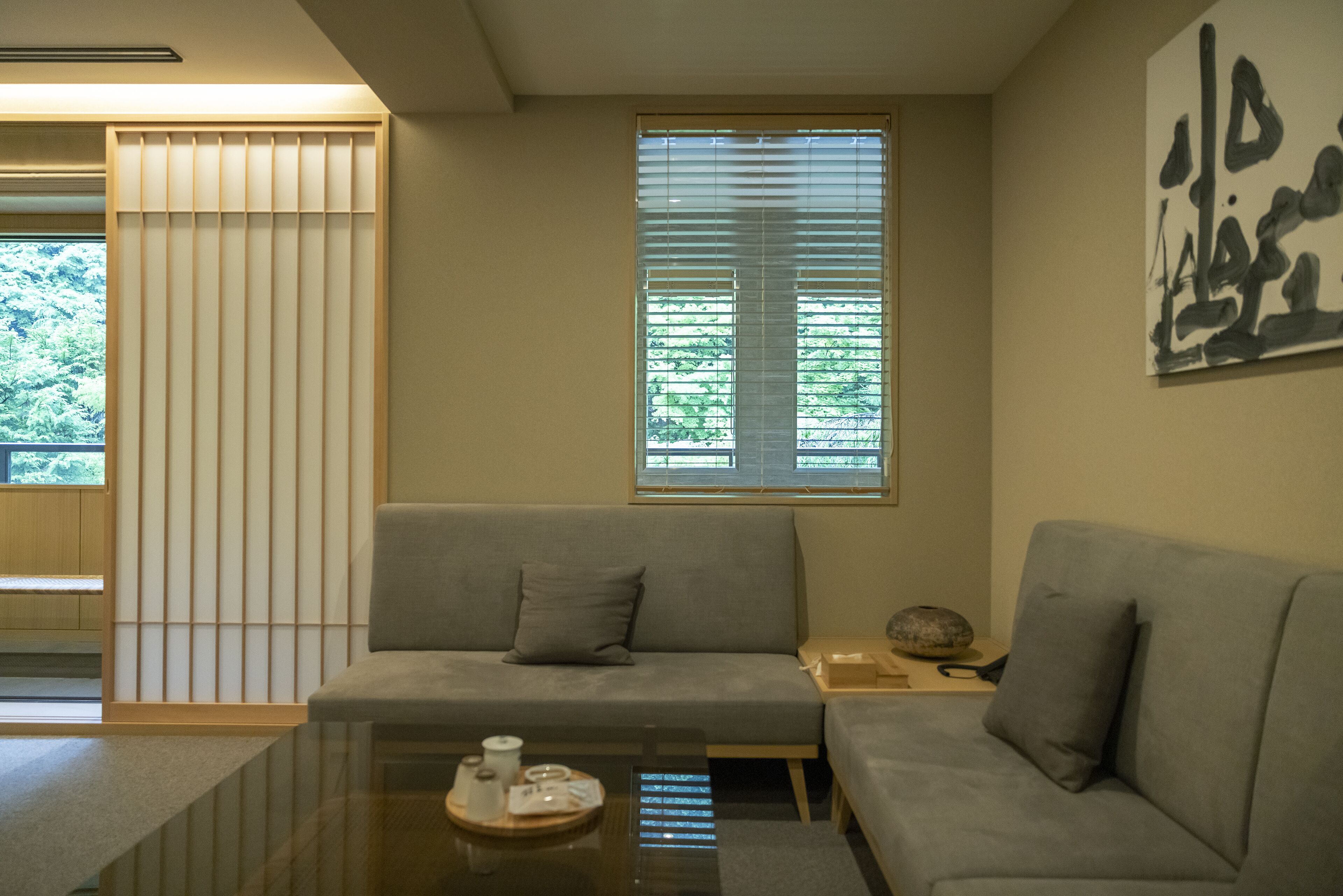 Ein modernes Wohnzimmer mit einem grauen Sofa, einem Glastisch und Blick auf Grün hinter den Jalousien.