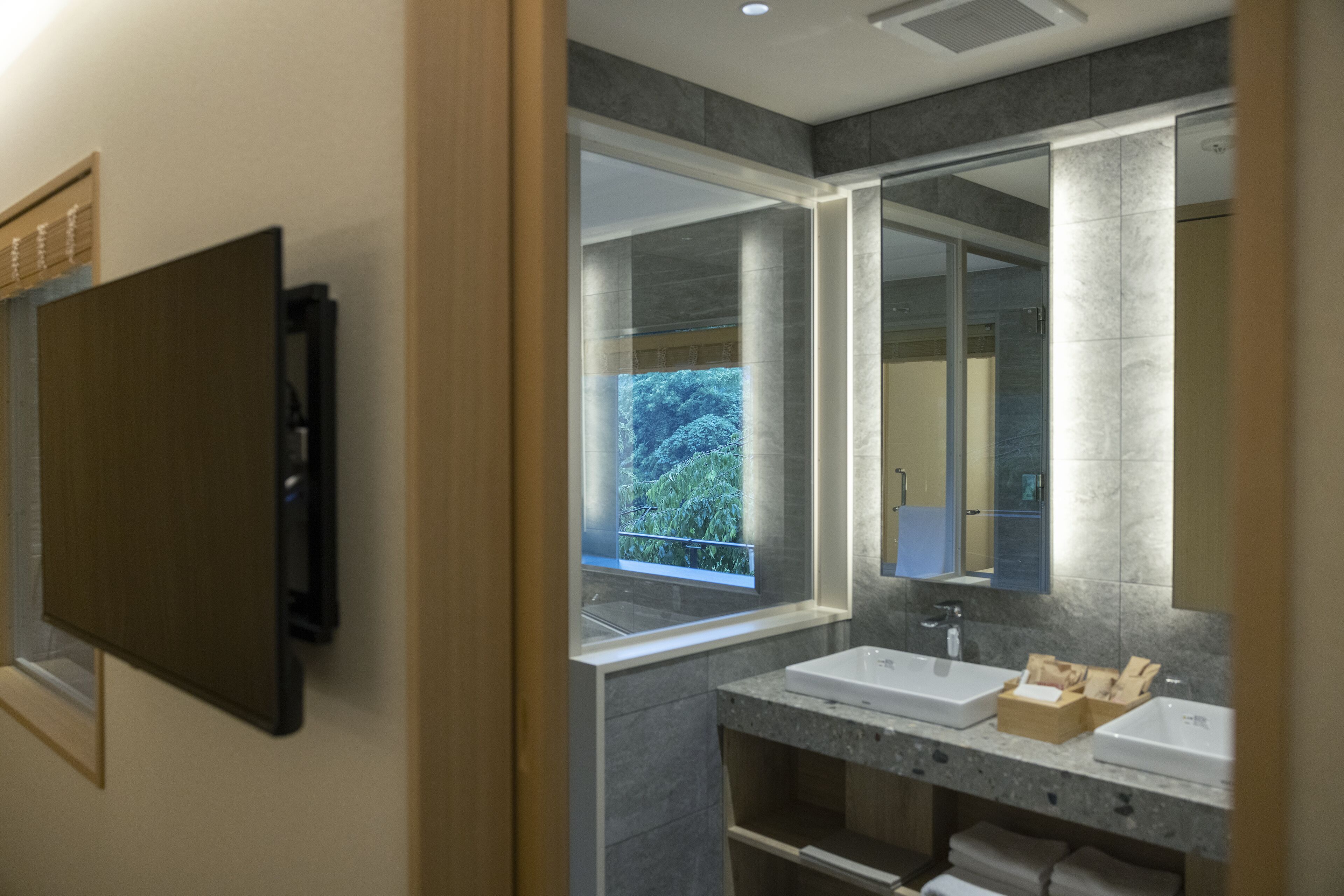 A modern bathroom with two sinks, a large mirror, and a window with a view of greenery.