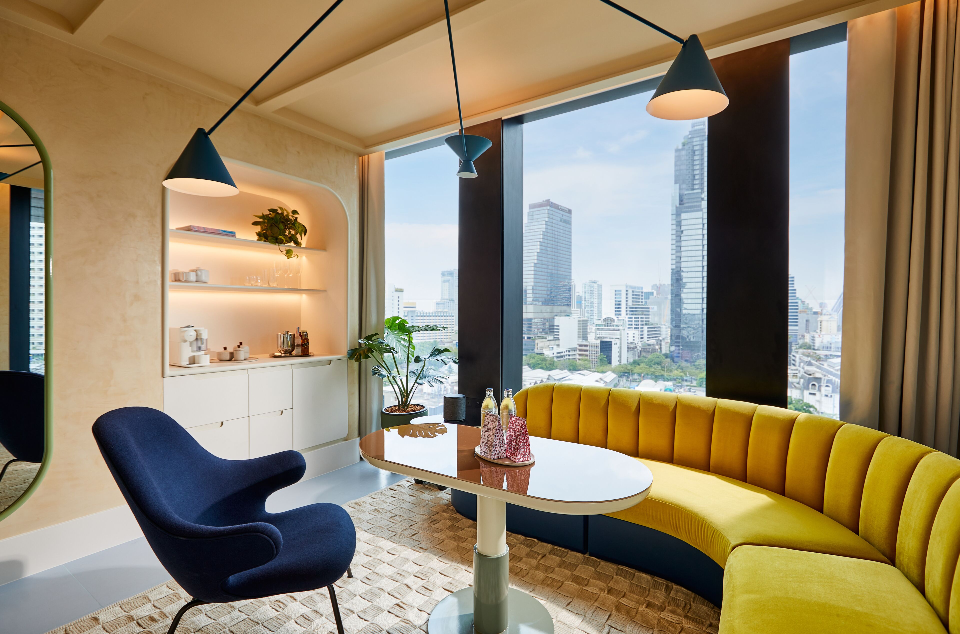 Una sala de estar moderna con un sofá amarillo, un sillón azul, una mesa de vidrio y vistas a la ciudad a través de amplios ventanales.