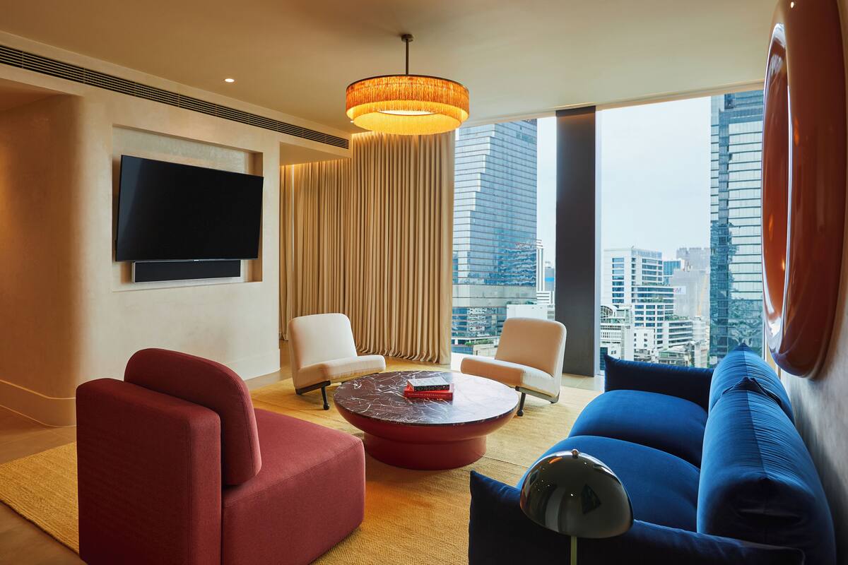 Ein modernes Hotelzimmer mit großem Fernseher, zwei Sesseln, einem roten Sofa, einem blauen Sofa, einem runden Couchtisch und einem großen Fenster mit Blick auf die Stadt.