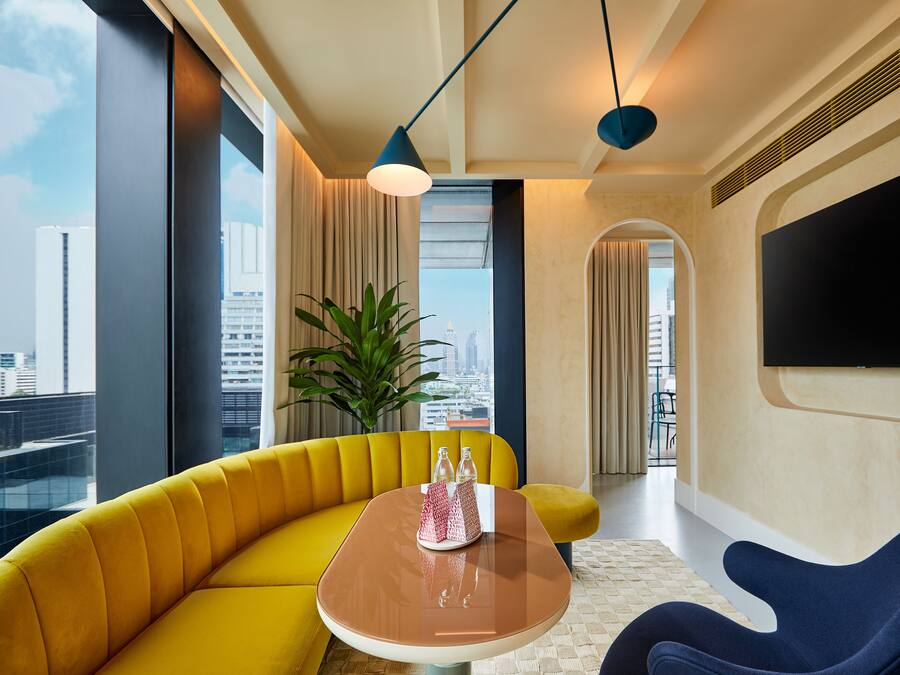 A modern living room with a yellow sofa, a brown table, and a blue chair. There is a large window with a city view, a potted plant, and a flat-screen TV mounted on the wall.