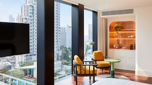 Une chambre d’hôtel moderne dotée d’une grande fenêtre, d’un téléviseur à écran plat, d’un lit, d’une petite table, de deux chaises jaunes et d’une étagère intégrée où sont disposés des objets décoratifs.