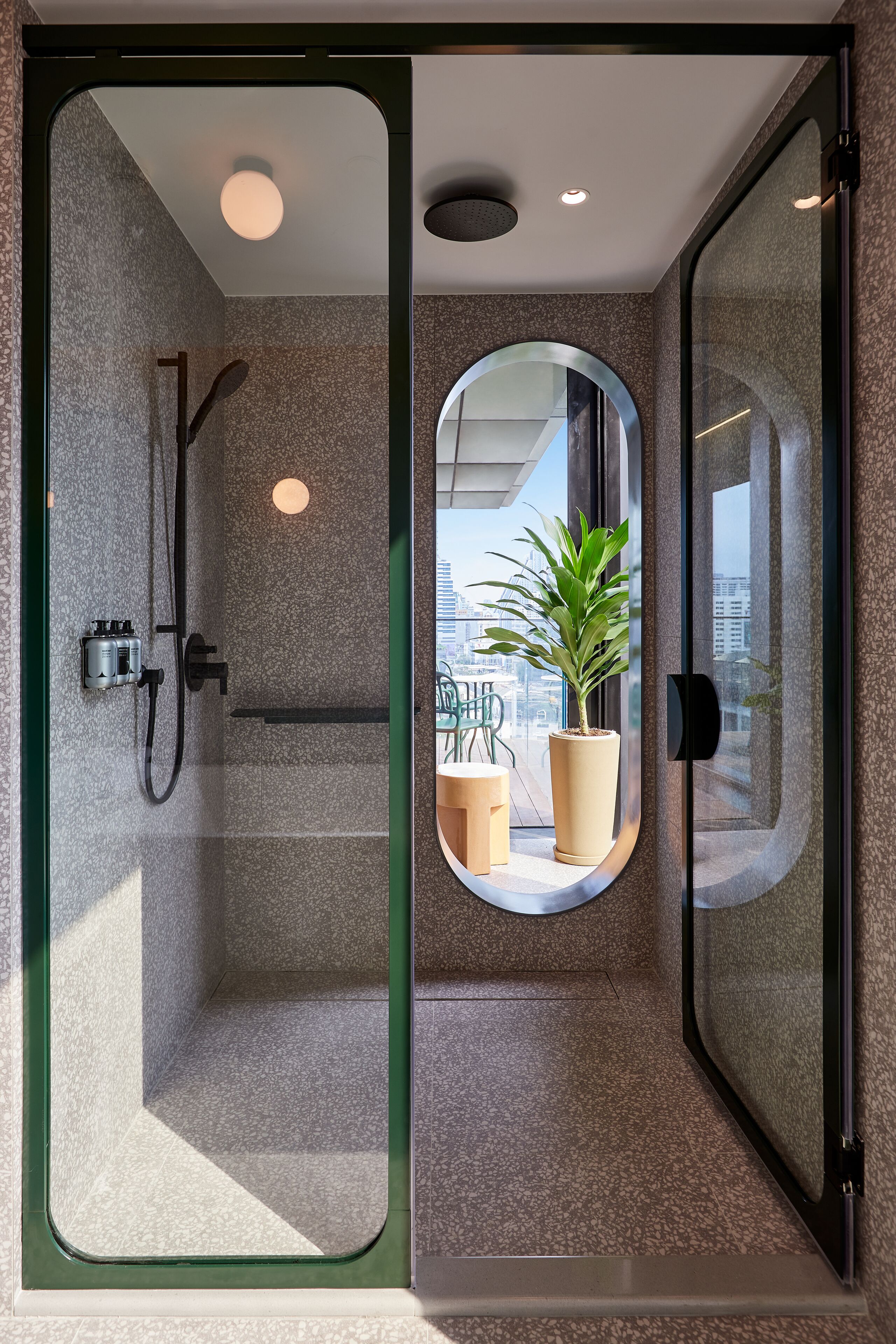 A modern bathroom with a glass-enclosed shower, a potted plant, and a view of the city.