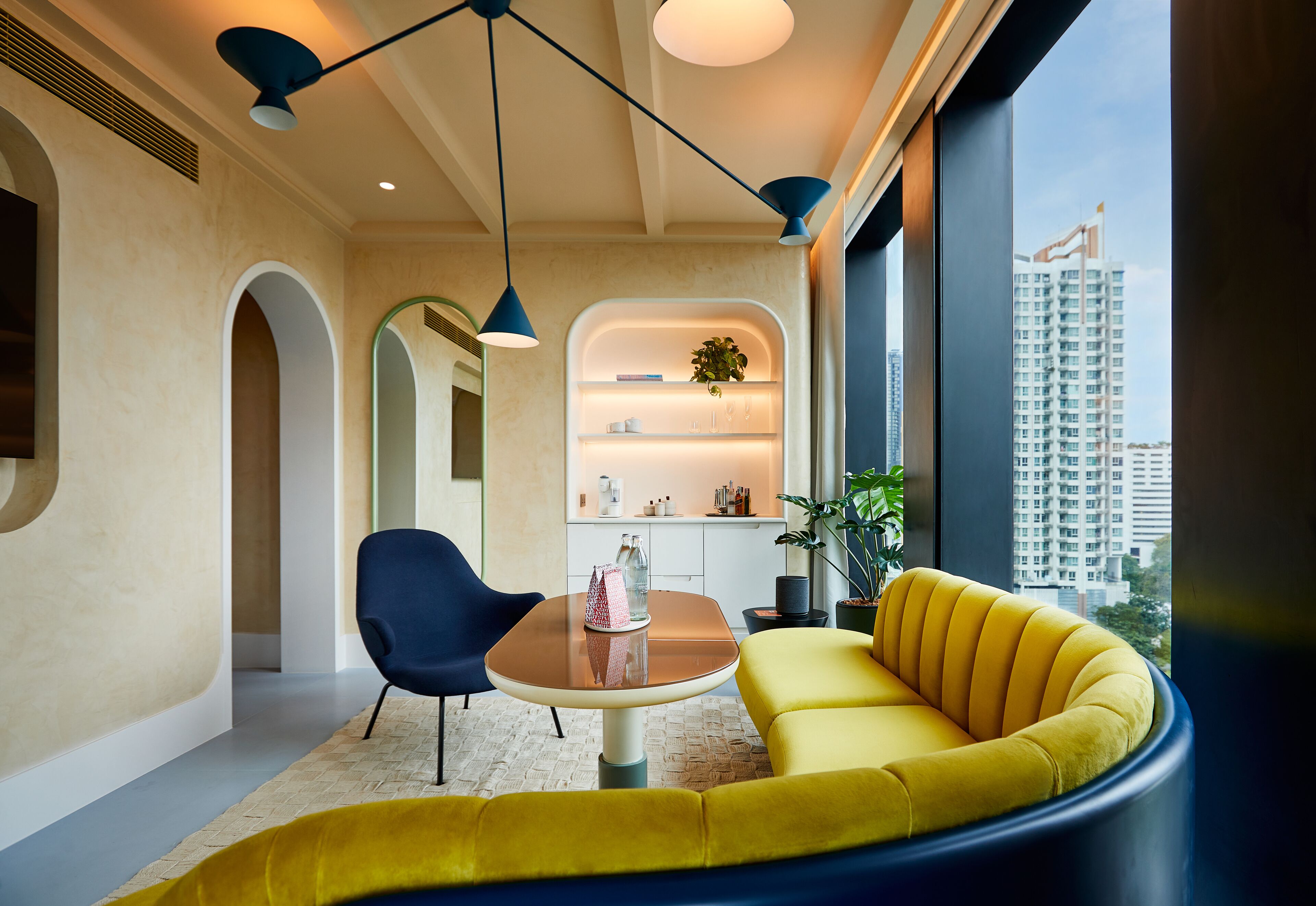 A modern interior with a yellow sofa, a round table, and blue chairs. There is a view of a cityscape through large windows.