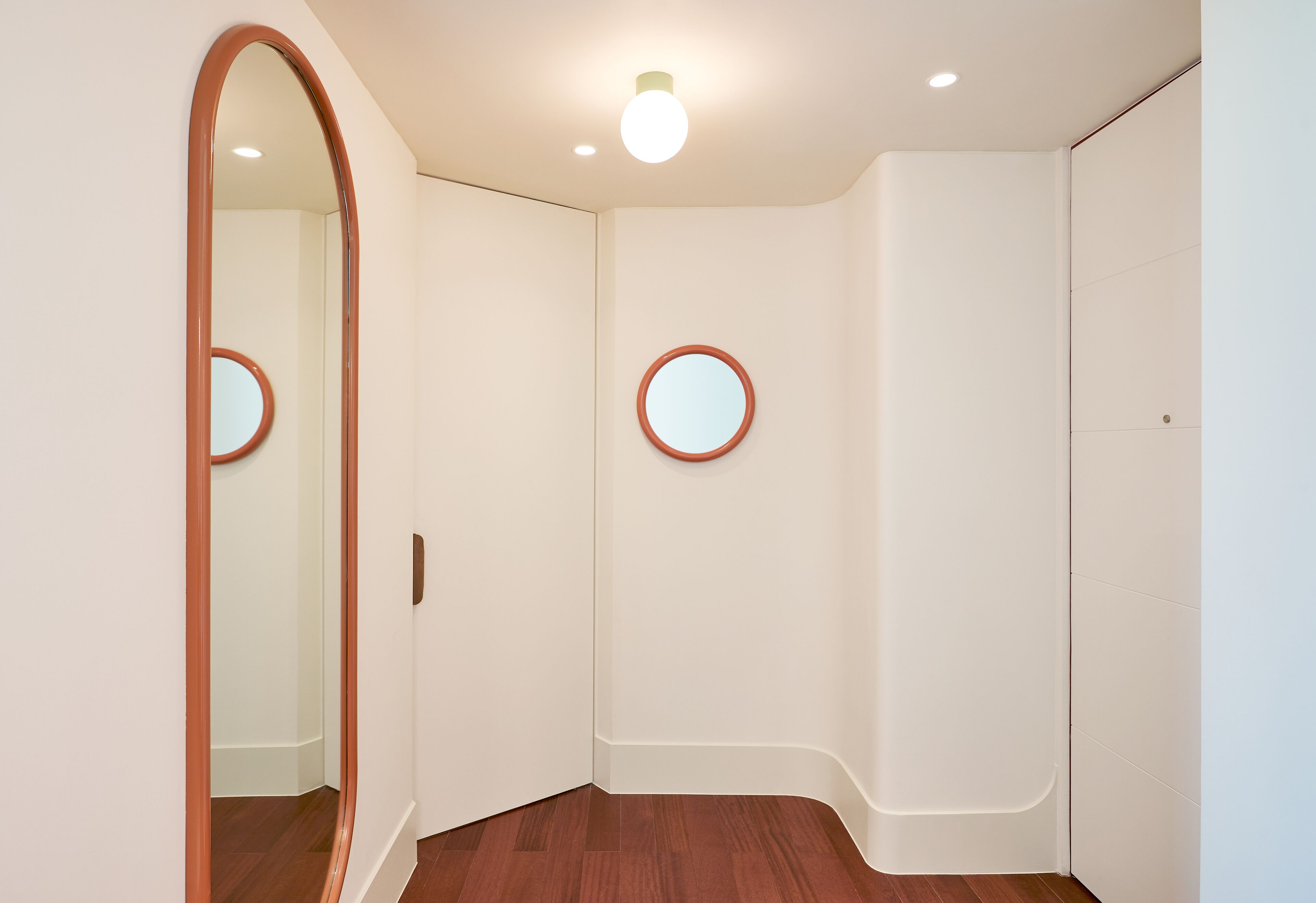 A room with a wooden floor, white walls, and a large mirror with a round frame.
