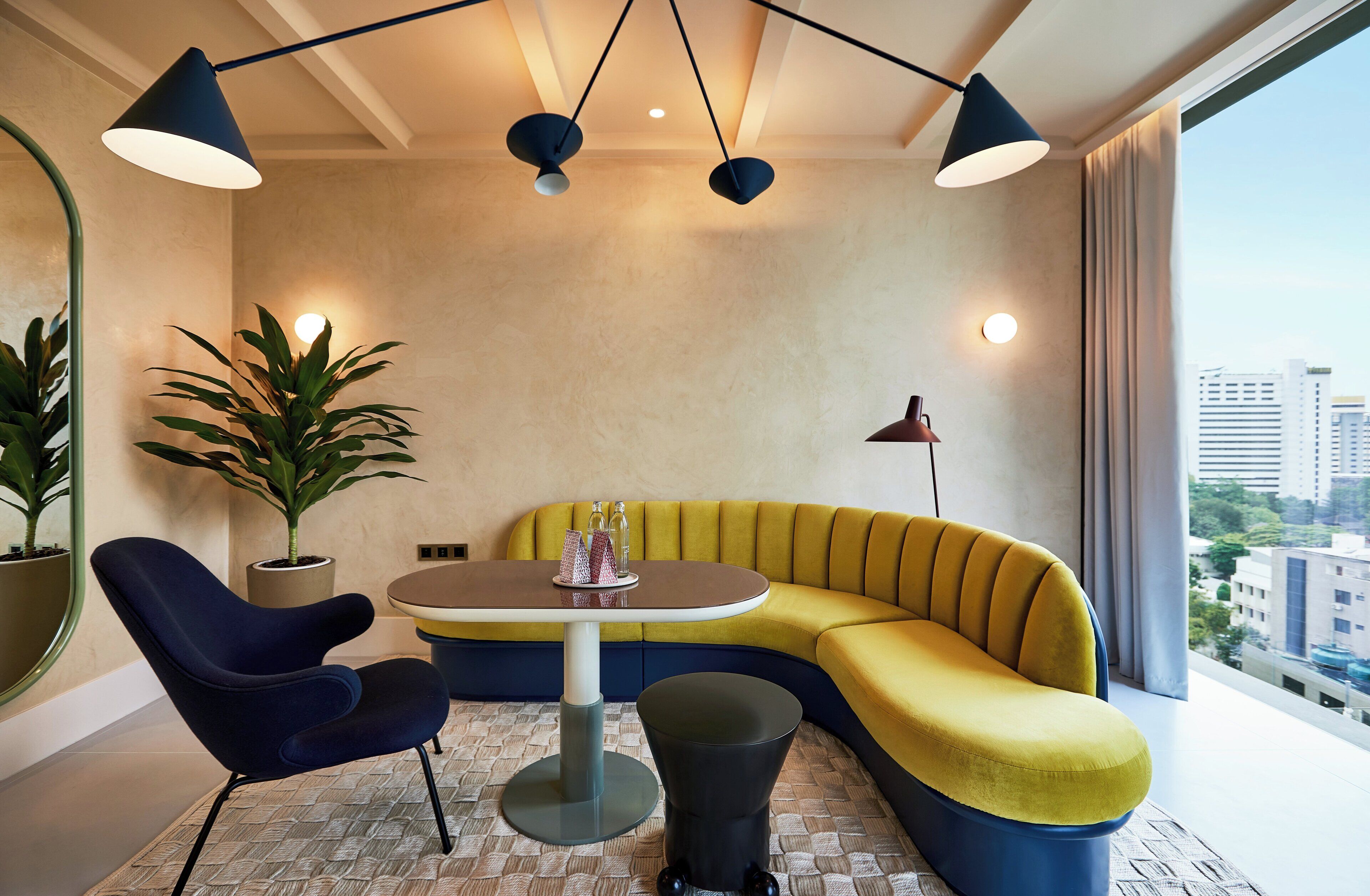 A modern room with a yellow sofa, a round table, and two chairs. There are two potted plants and a floor lamp.
