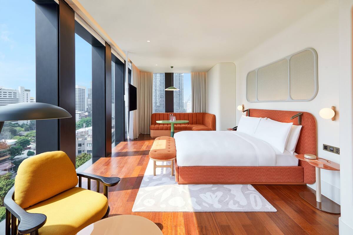 Ein modernes Hotelzimmer mit einem großen Bett, einem gelben Sessel, einem kleinen Tisch und Blick auf die Stadt durch große Fenster.
