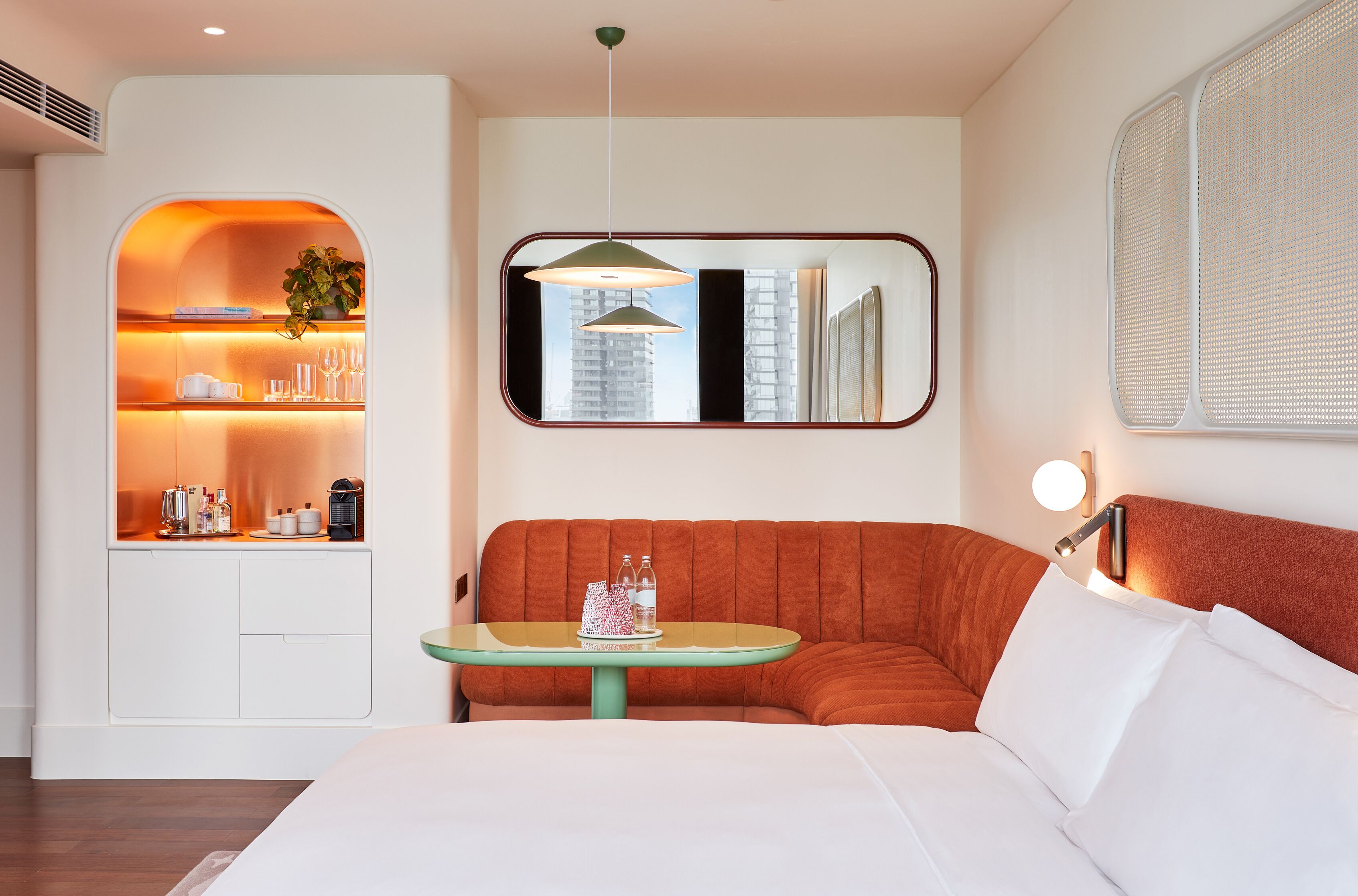 A modern hotel room with a bed, a seating area with a table, a built-in kitchen, and a large window.