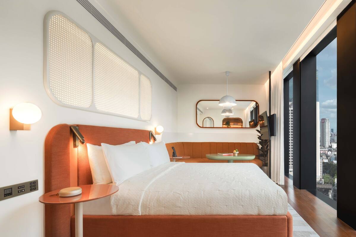 Ein modernes Hotelzimmer mit einem großen Bett, einem Nachttisch, einem runden Tisch, einem Spiegel und Blick auf die Stadt durch große Fenster.