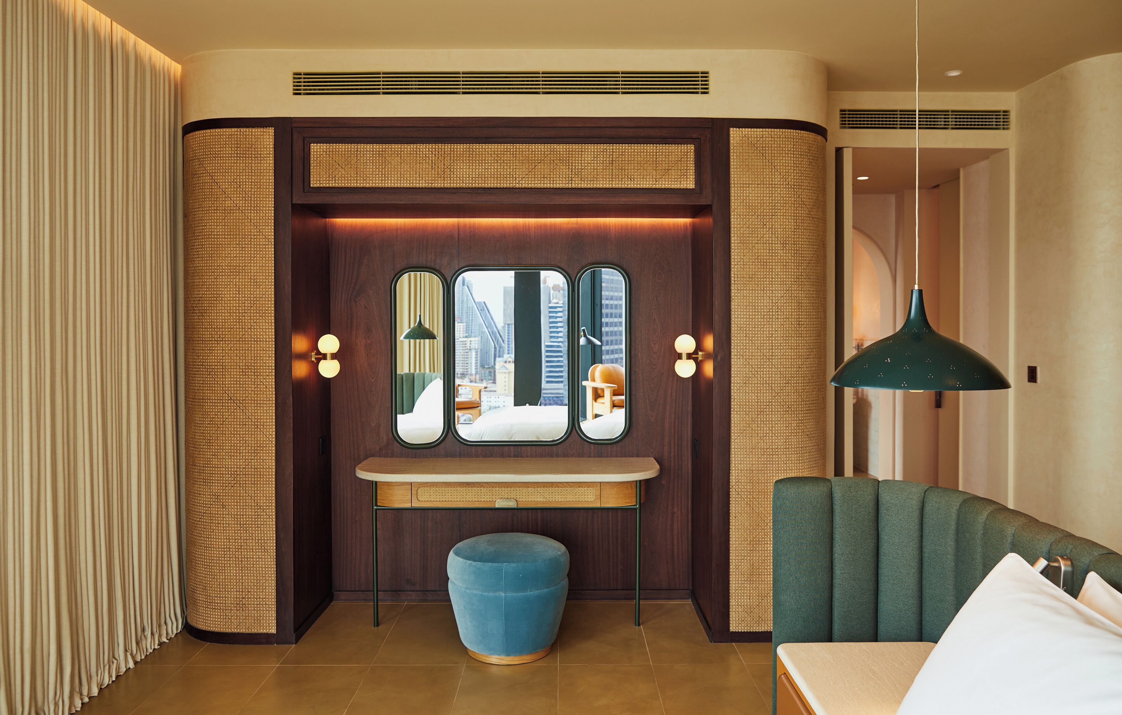 A hotel room with a large mirror, a vanity with lights, a blue ottoman, and a green sofa.