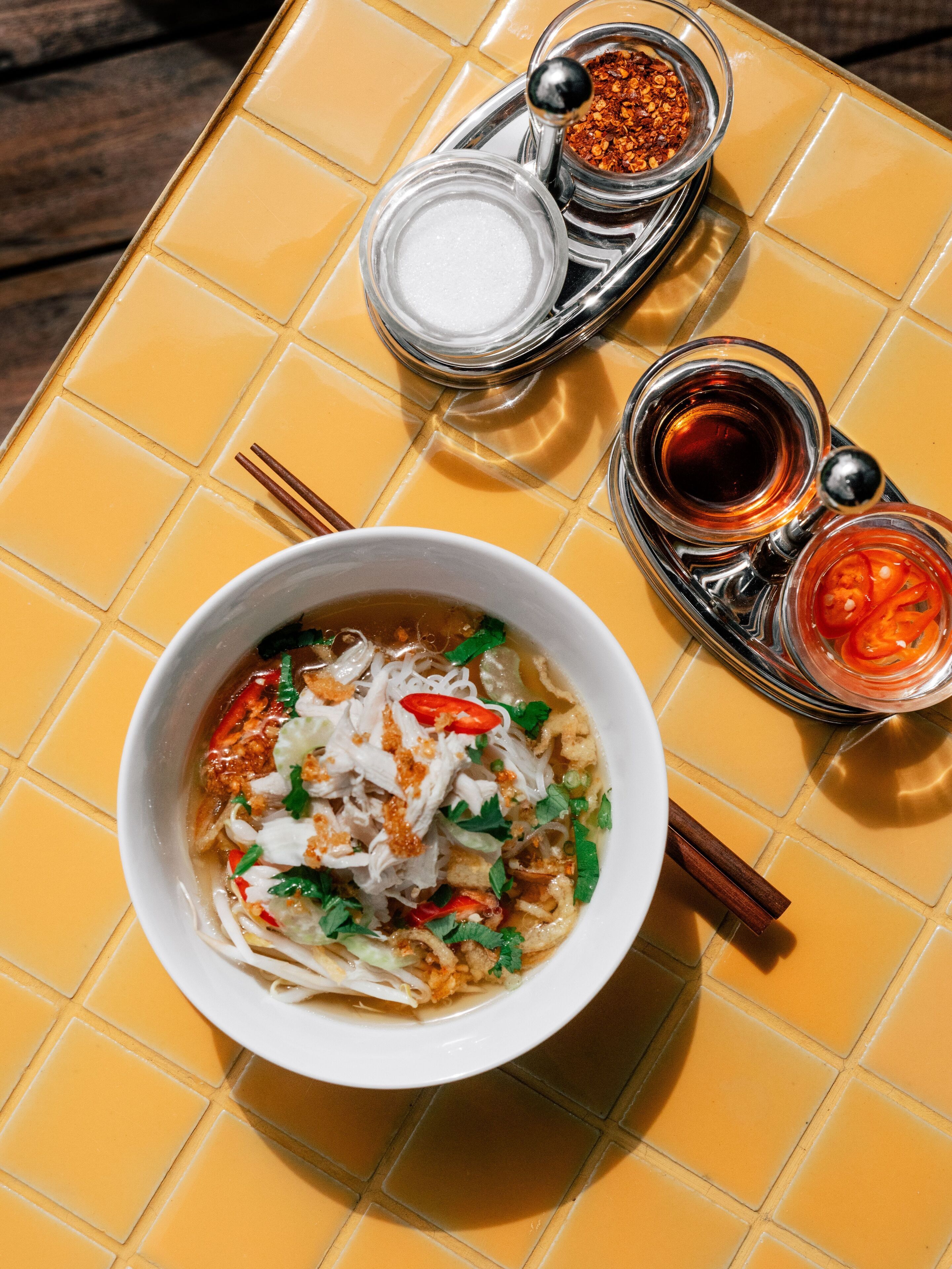 A bowl of noodle soup with toppings, a small bowl of chili, and a container of soy sauce on a yellow tiled table.