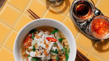 A bowl of noodle soup with toppings, a small bowl of chili, and a container of soy sauce on a yellow tiled table.