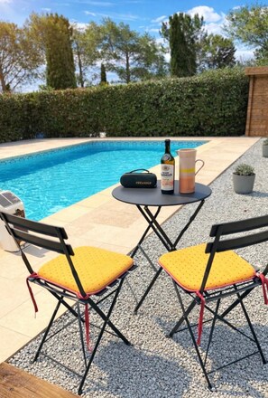 A poolside area with two yellow cushioned chairs, a table with a bottle and a pitcher, and a small black bag.