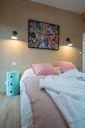 Une chambre à coucher comprenant un lit, une œuvre d’art colorée accrochée au mur, deux lampes de chevet et une petite table de chevet turquoise avec des tiroirs.