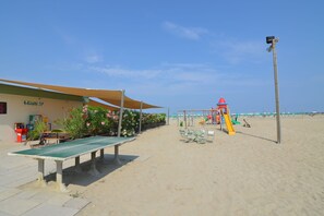 A beachside area with a shaded table and chairs, a children's play structure, and a building with a sign. - Relaxing Resort Apartment (Lido di Spina)