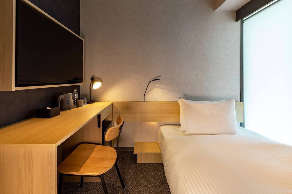 Quarto de hotel moderno com escrivaninha de madeira, uma cadeira, cama com roupa de cama branca, televisão de tela plana na parede e mesa de cabeceira com abajur.