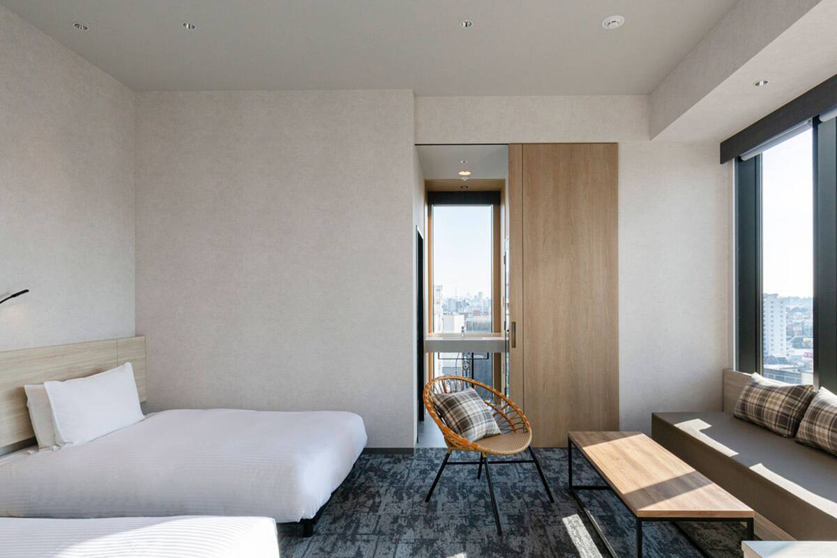 Quarto de hotel moderno com uma cama grande, área de estar com um banco e uma cadeira, e vista da cidade através de uma janela ampla.