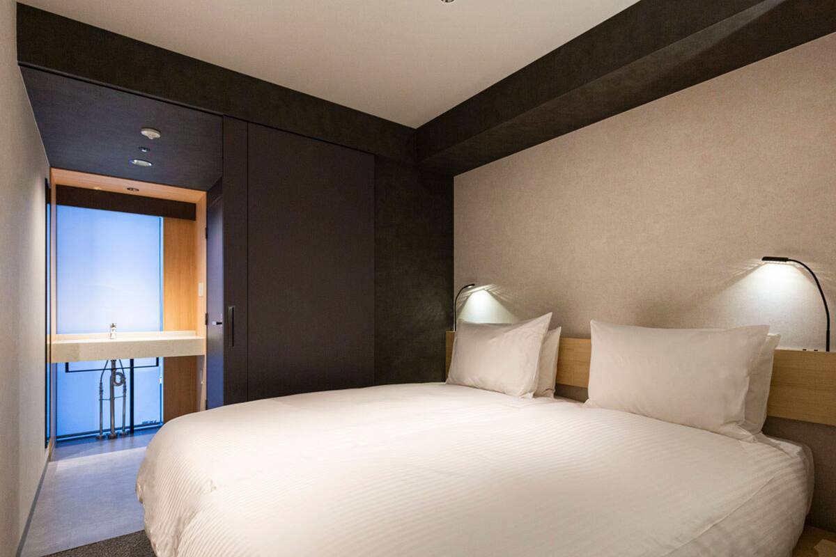 Quarto de hotel moderno com uma cama grande, duas luminárias de cabeceira, um painel de madeira na cabeceira e vista para o banheiro.