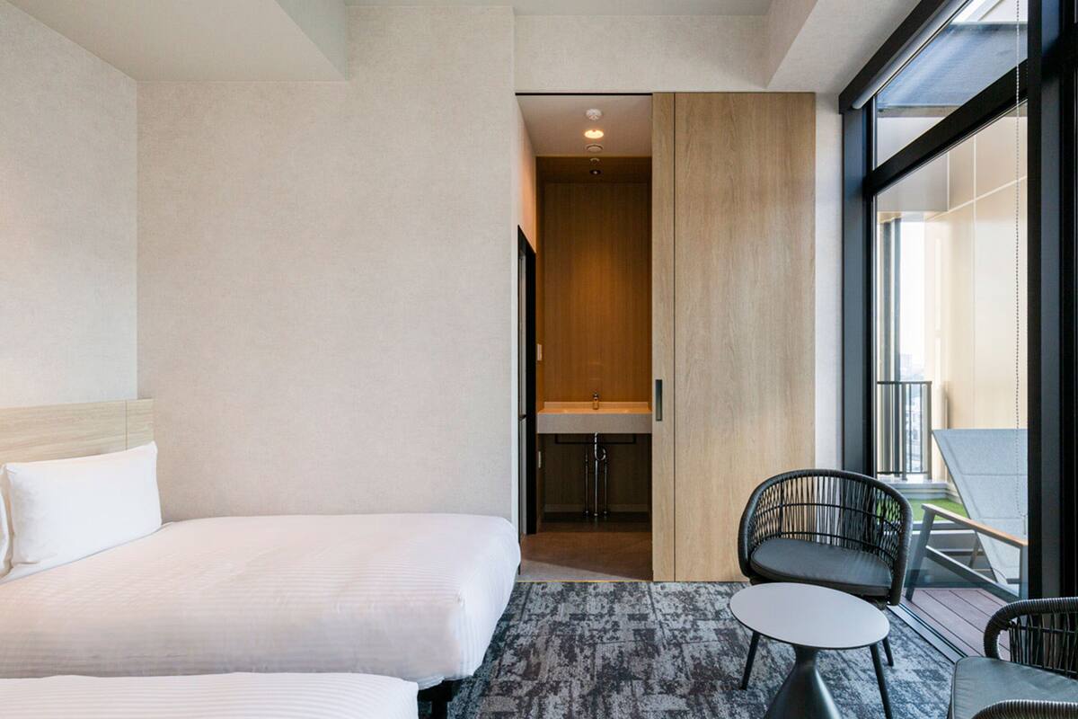 Quarto de hotel com duas camas, uma pequena área de estar com cadeiras e vista para o exterior.