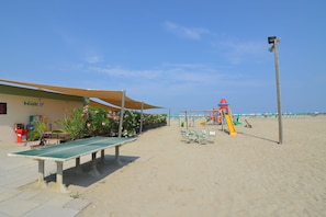 Un espace aménagé au bord de la plage, avec une table et des chaises à l’ombre, une aire de jeux pour enfants et un bâtiment portant une enseigne.