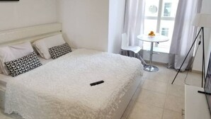 A neatly made bed with a fluffy white cover, a small bedside table with a lamp, and a window with curtains. - Luxury Private Room in an Apartment Next to Barasti Beach  (Dubai)