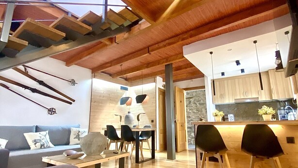 En moderne stue med træloft, et spiseområde og et køkken, der kan ses gennem en åben døråbning.
