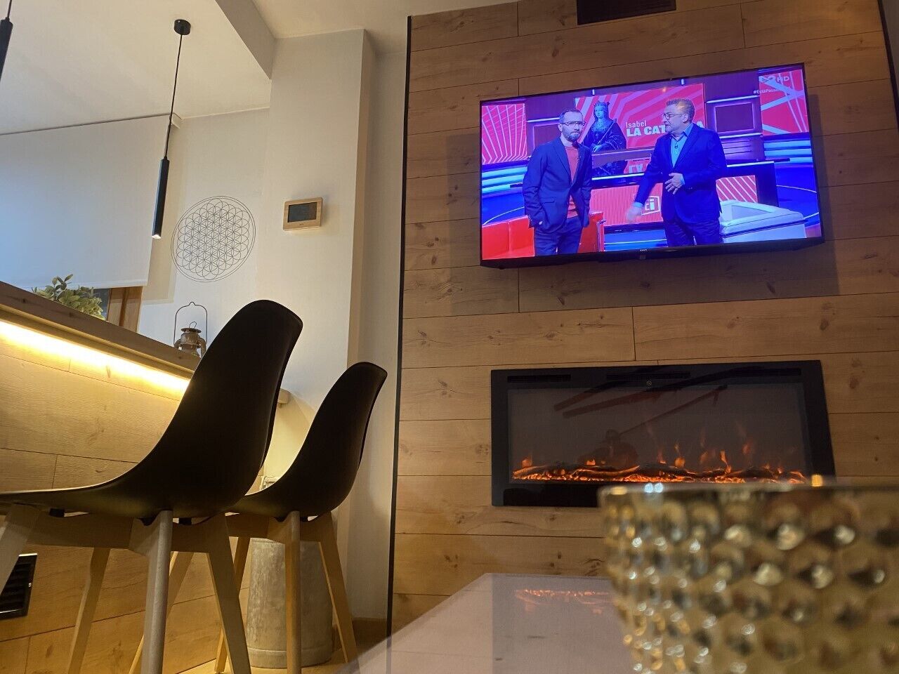 A modern living room with a flat-screen TV mounted on the wall, a fireplace below it, and two black chairs in the foreground.