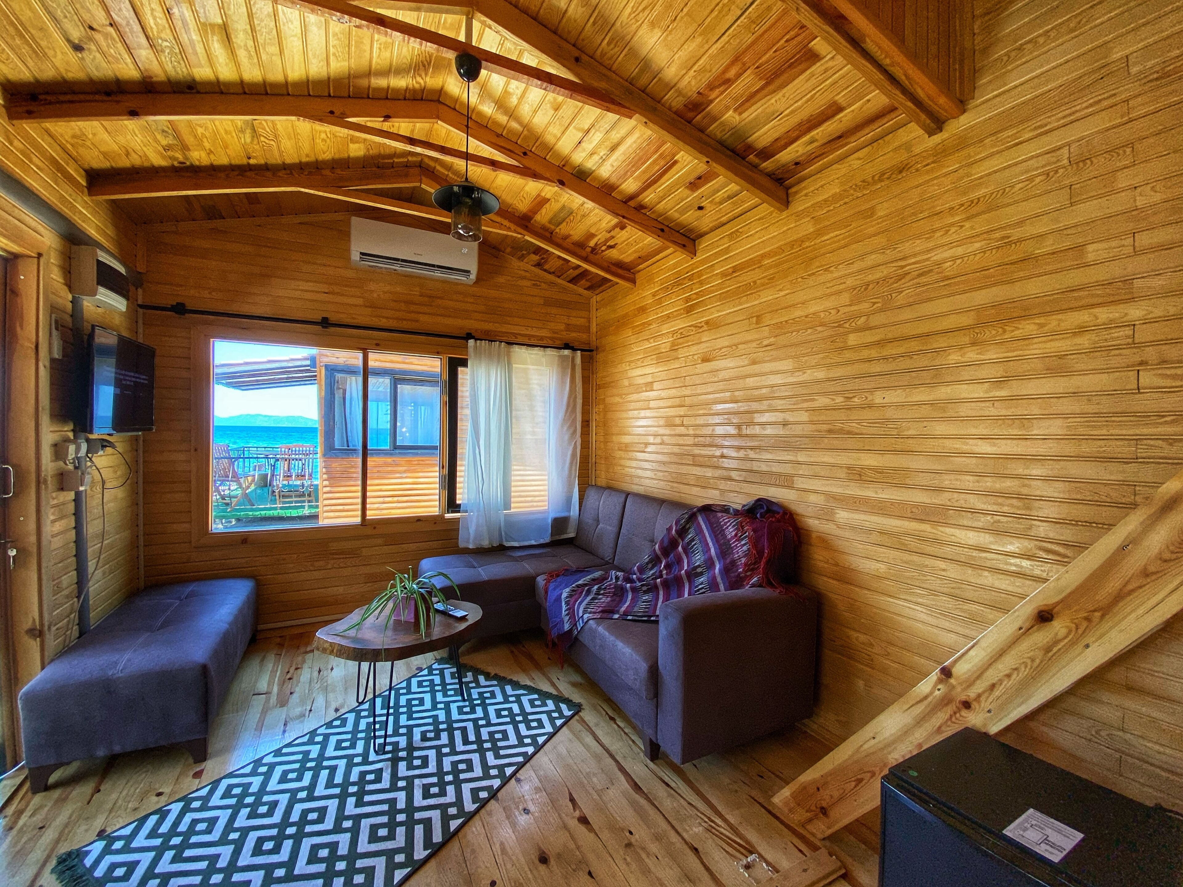 Interior acogedor de una cabaña con techo de madera, un sofá, una mesita con una planta y vista al océano a través de un ventanal grande.