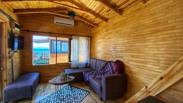 Interior acogedor de una cabaña con techo de madera, un sofá, una mesita con una planta y vista al océano a través de un ventanal grande.