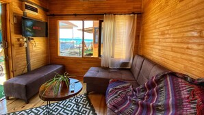 Interior acogedor de una cabaña con techo de madera, televisor montado en la pared, un sofá, una mesita con una planta y vista a la playa a través de una ventana.