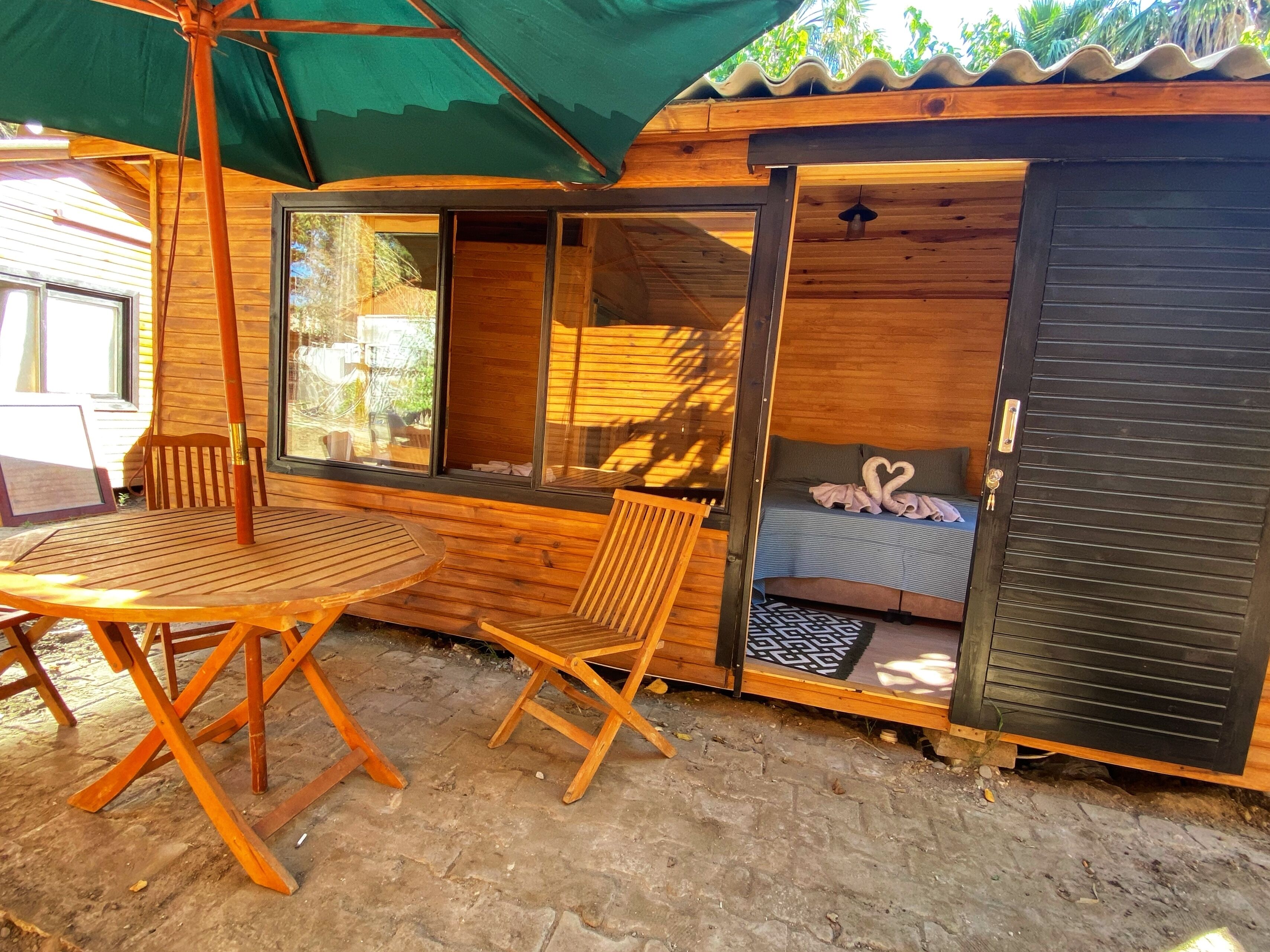 Uma cabana de madeira com porta deslizante, uma cama com almofadas em formato de coração, uma mesa e uma cadeira.