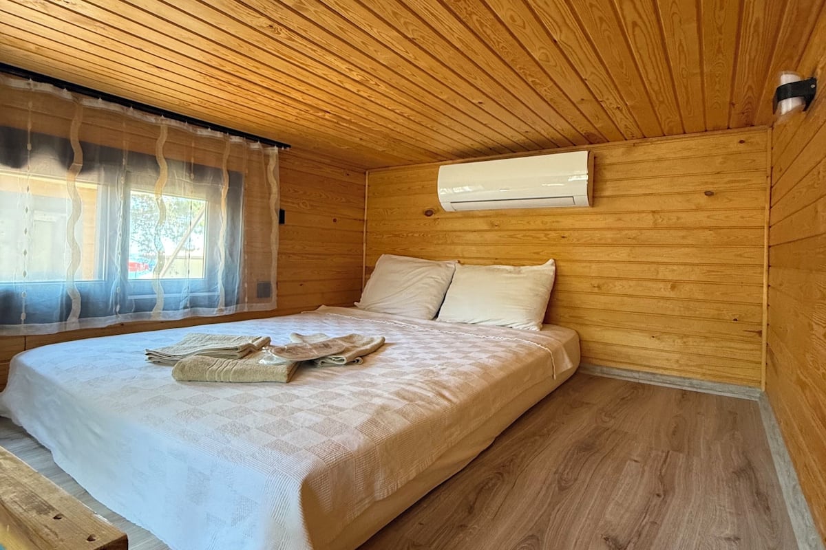 A wooden cabin bedroom with a bed, a window with curtains, and an air conditioning unit.