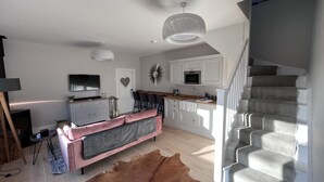 A modern kitchen with a dining area, a staircase, and a living room with a sofa and TV.