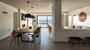 Un comedor moderno con una mesa larga y sillas, una lámpara en el techo y vista al mar a través de amplias ventanas.