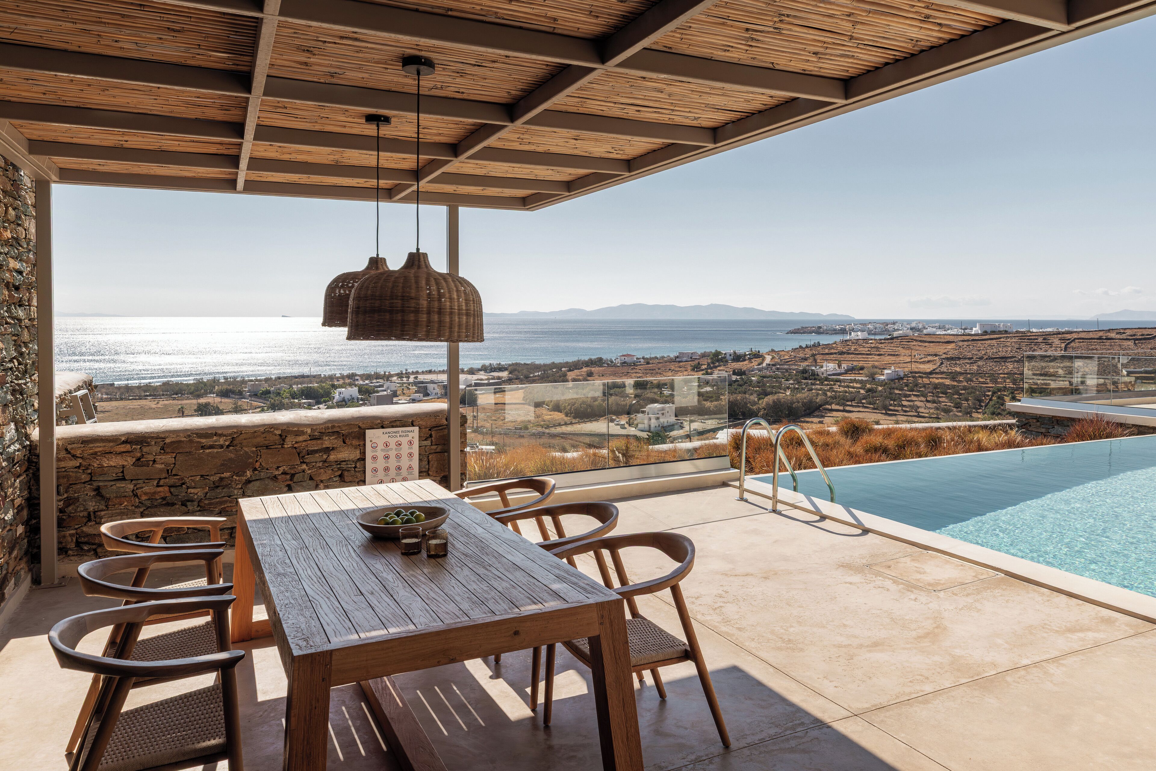 A wooden table and chairs under a pergola with a view of a swimming pool and the sea.