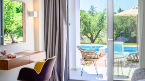 A modern hotel room with a wooden desk, a purple chair, a bed, and a sliding glass door leading to an outdoor area with a pool and lounge chairs. - Alonaki Resort (Preveza)
