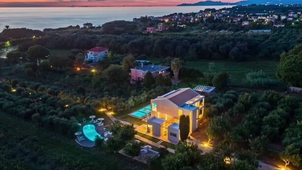 An aerial view of a modern house with a swimming pool, surrounded by lush greenery and a view of the sea at dusk. - Alonaki Resort (Preveza)